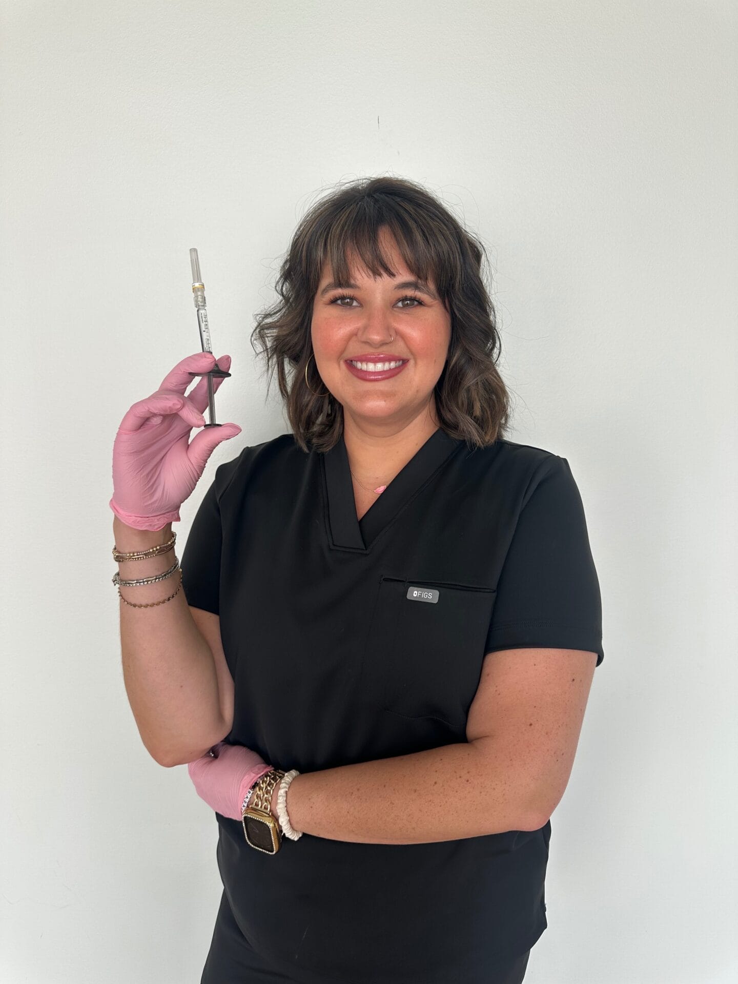 A woman in black scrubs and pink gloves holds a syringe upright, smiling at the camera against a plain white background.