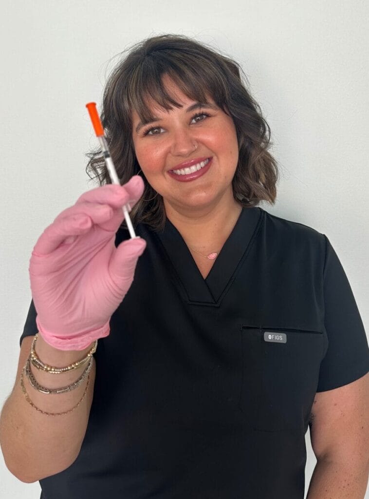 A woman in black scrubs and pink gloves holds up a syringe and smiles at the camera against a plain white background.