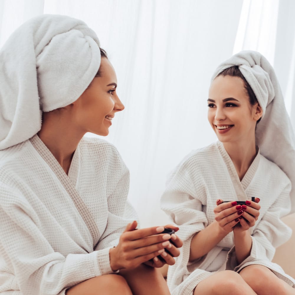 Two women in white robes receiving facials on a couch in a spa.