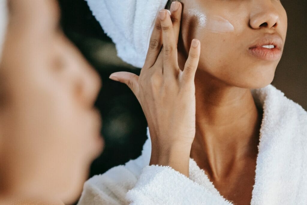 close up of brown-skinned woman in white robe and headtowel applying moisturizer to her cheek