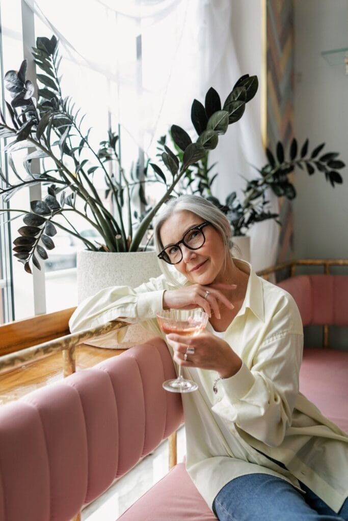 An older woman in glasses is sitting on a pink couch with a glass of wine, showcasing the results of a proactive anti-aging skincare routine including cosmetic injectables.