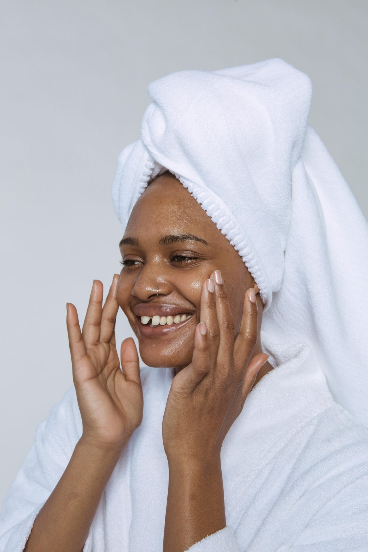 A woman in a white towel is gently applying lotion to her face after a skin treatment.