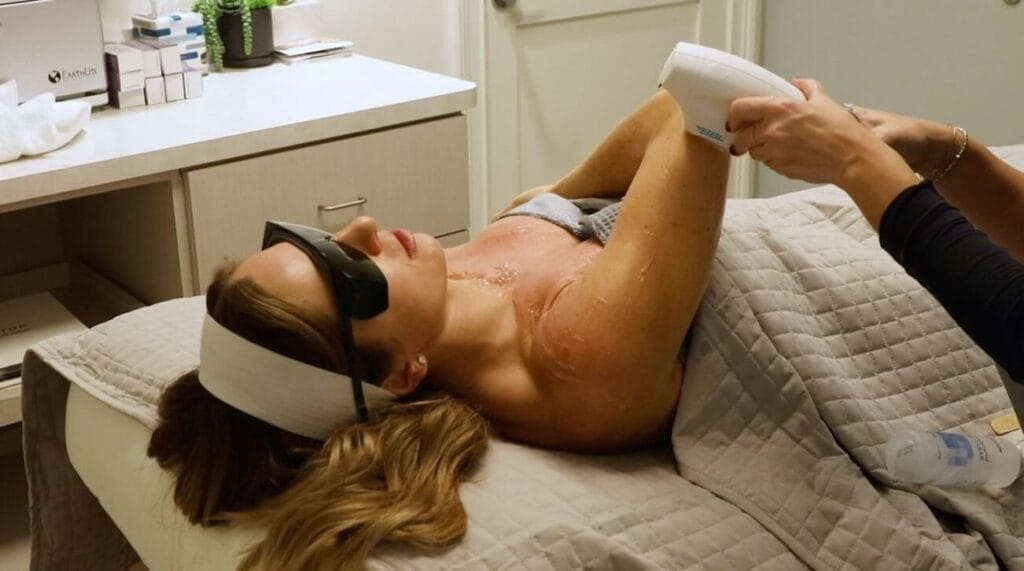 Person receiving laser hair removal on their underarm while lying on a treatment table, wearing protective sunglasses and a headband.