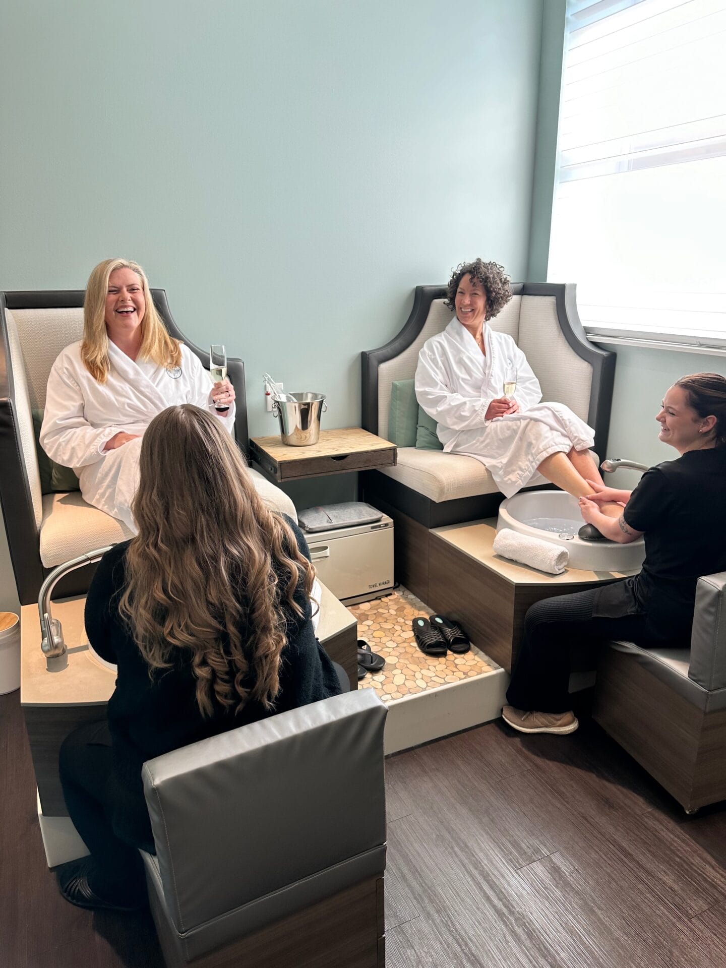 Two women in white robes sit in spa chairs receiving pedicures from two spa professionals. One woman on the left is smiling and holding a glass of champagne, enjoying her Platinum Day of Indulgence.