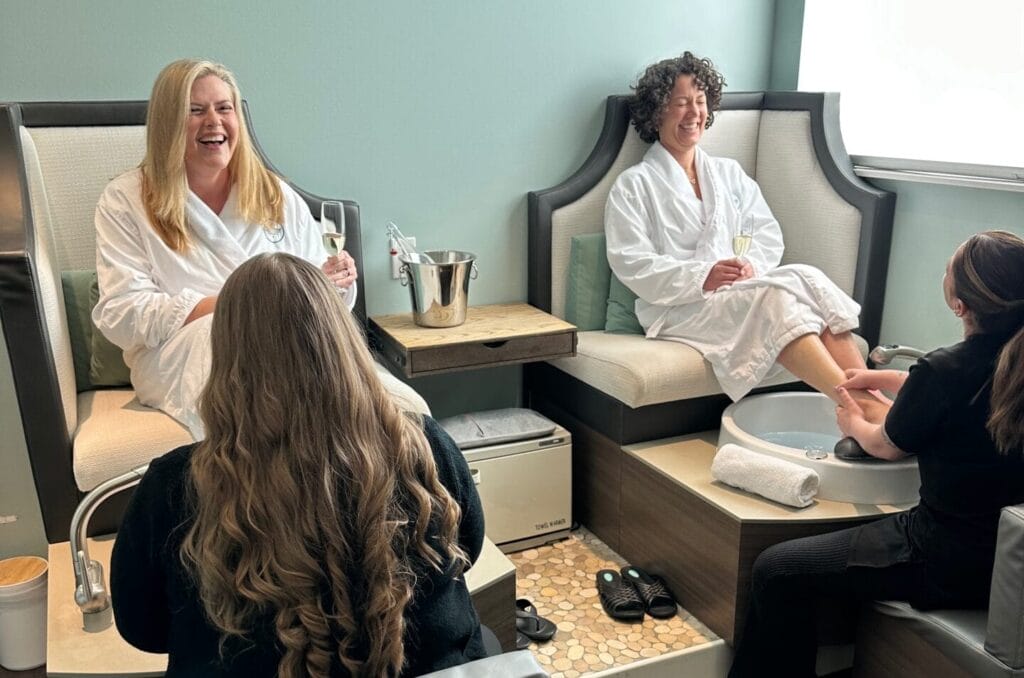 Two women wearing white robes are enjoying a Day of Indulgence with pedicures from salon staff in a spa setting. Seated on individual spa chairs and holding glasses of beverages, they revel in this platinum experience.