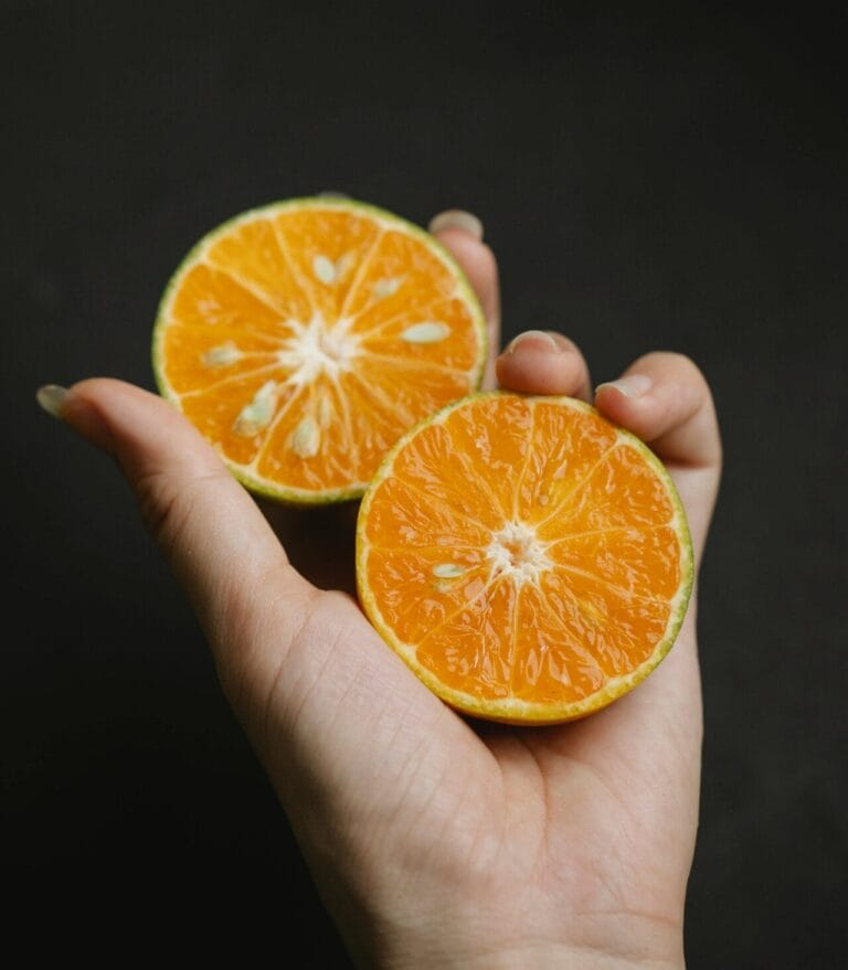 A hand holding two halves of an orange, rich in Vitamin C, against a dark background.