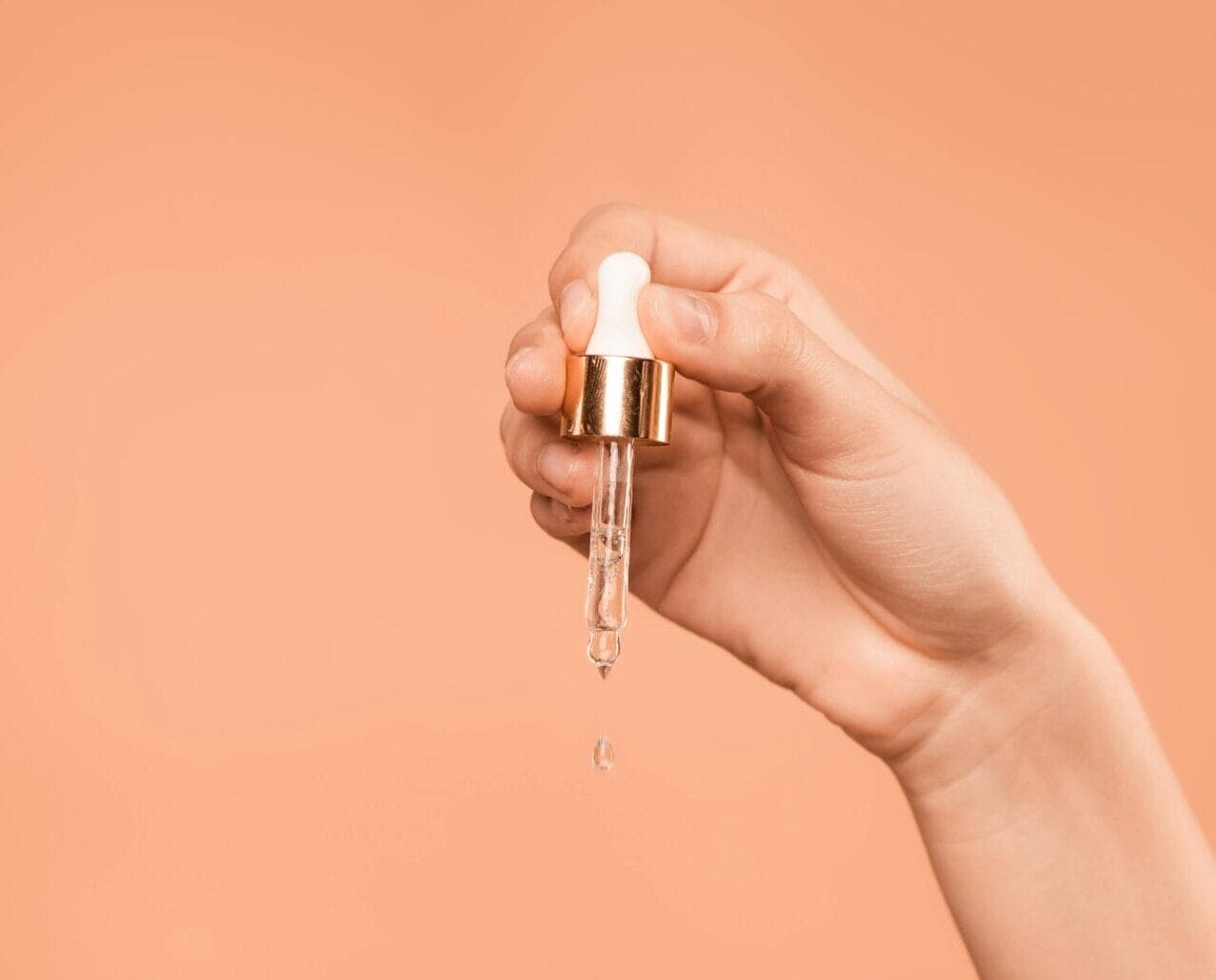 A hand holds a dropper above another hand, releasing a Vitamin C-infused drop of liquid against a peach-colored background, like skincare superheroes ready to save the day.