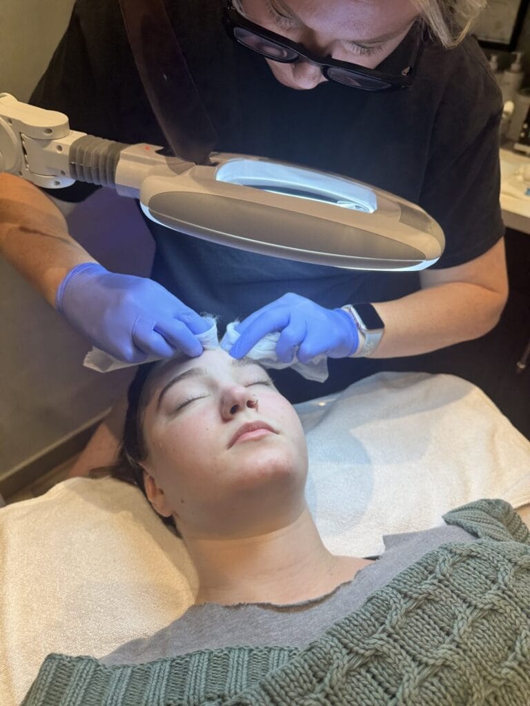 A teen is receiving a clinical facial from another individual wearing gloves and using a lighted magnifying lamp.