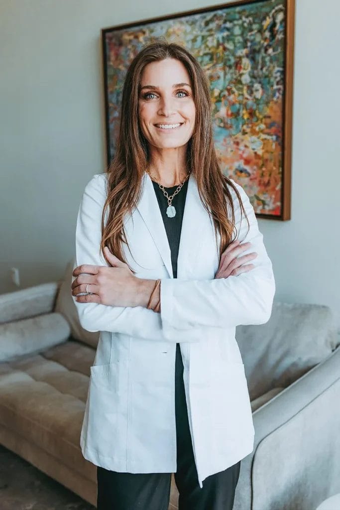 Tara Barton, featured in HealthScope Magazine, stands with arms crossed and smiling in front of a colorful painting and a gray couch, perfectly embodying the serene elegance of Center Medspa.