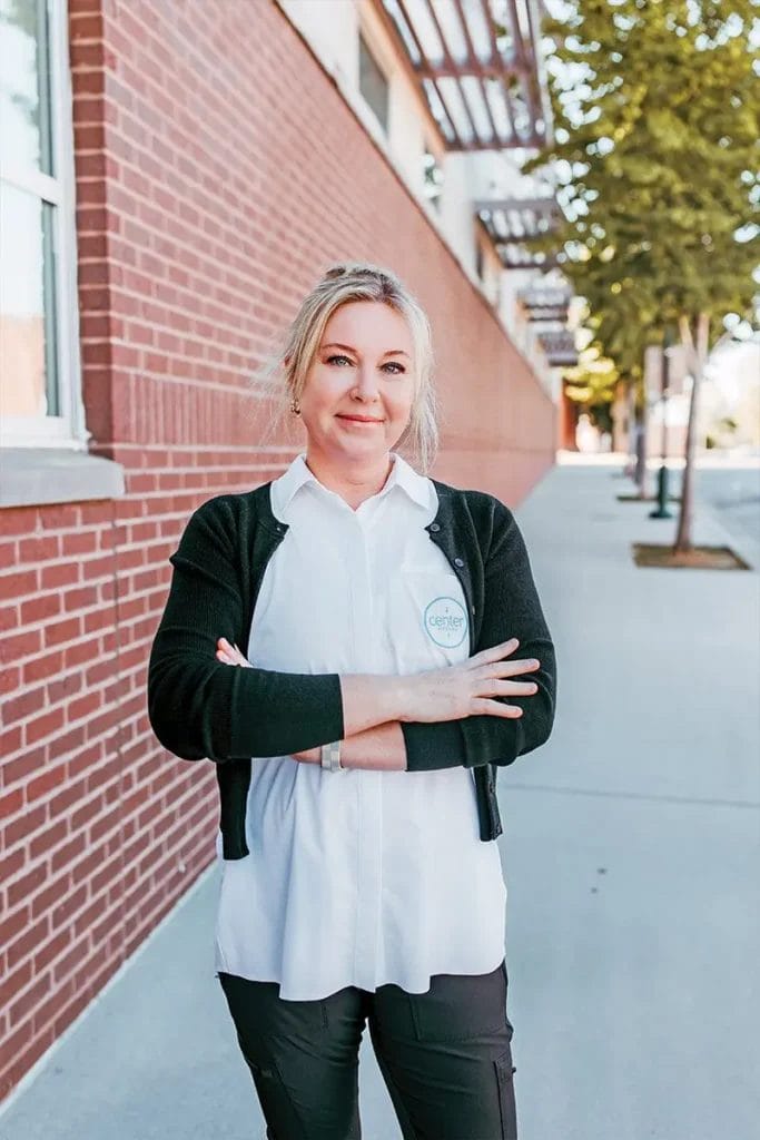 Daisy Tucker, with her blonde hair and confident stance, stands on the sidewalk beside a brick building. Dressed in a white shirt and black sweater, she could easily be featured in HealthScope Magazine for her poised presence.