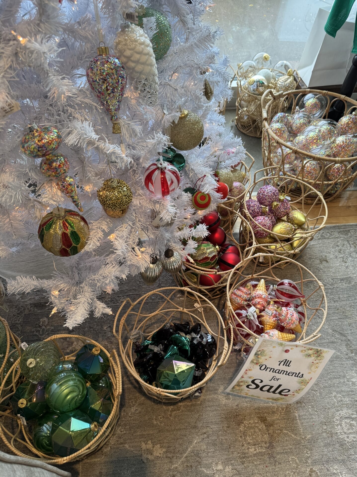 A beautiful white Christmas tree stands adorned with various ornaments in baskets nearby, perfect for your holiday shopping. A sign indicates all ornaments are on sale, making it an irresistible festive find.