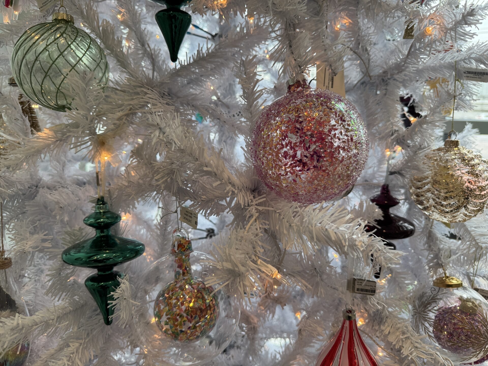 Close-up of a white Christmas tree decorated with assorted glass ornaments, including a large pink glittery bauble, green and red shapes, and small twinkling lights—perfect for setting the festive mood during your 12 Days of Christmas holiday shopping spree.