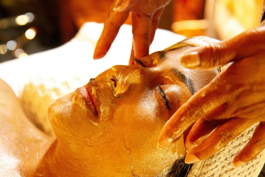 A person receiving a soothing facial treatment, with a golden mask enhancing their glow, lies peacefully on a spa bed while expert hands gently press on their skin, indulging in luxurious skincare.