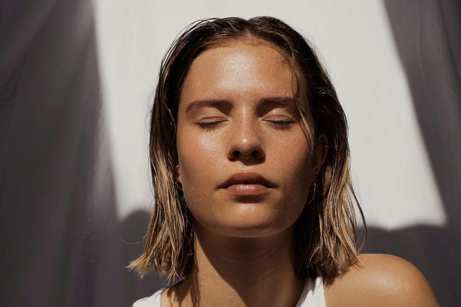 A person with short hair and closed eyes basks in sunlight with a serene expression, as if touched by light and laser magic, set against a blurred background.