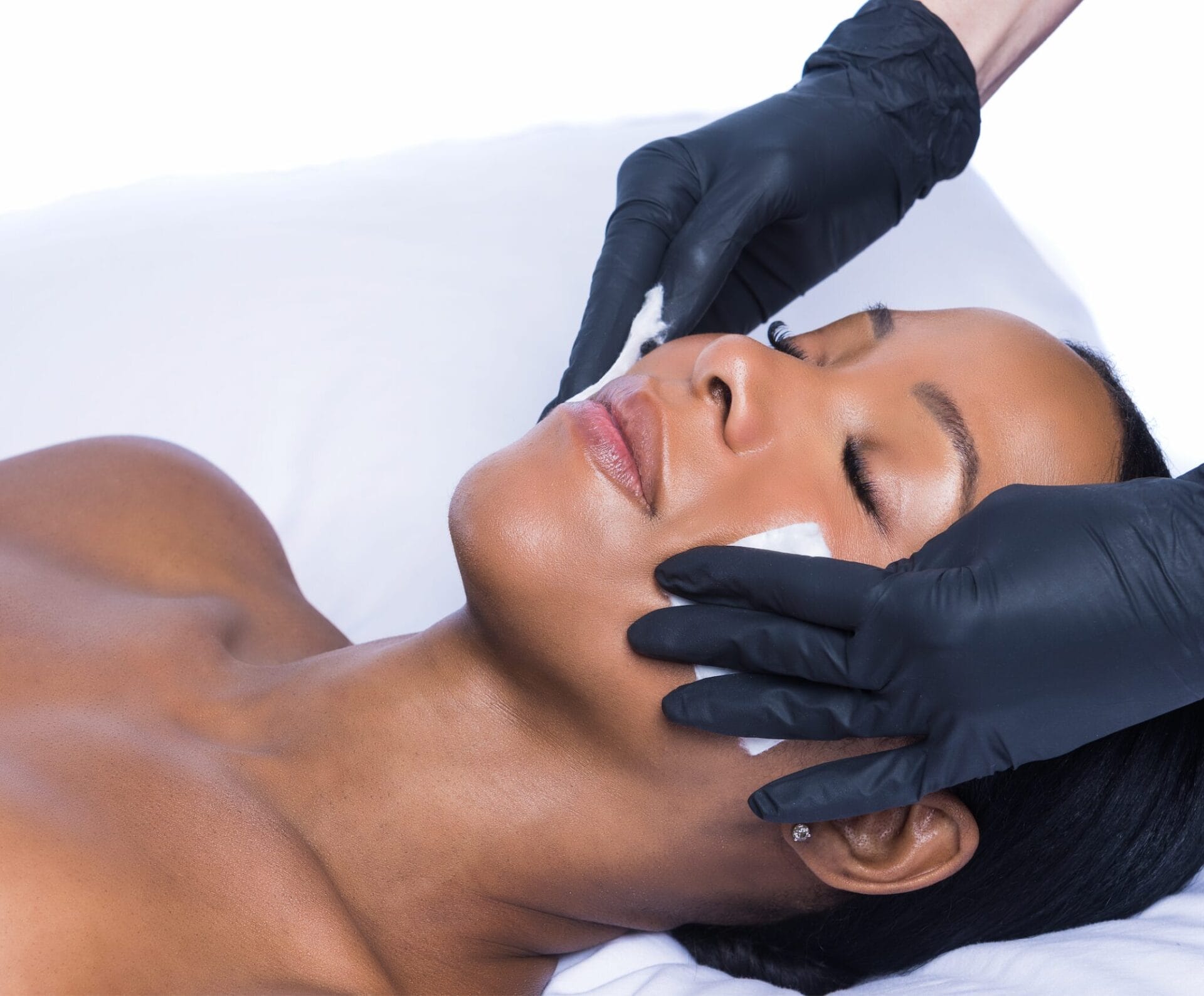 At Center MedSpa, a person lies peacefully with eyes closed, receiving a soothing skincare treatment. Gloved hands gently press pads on their cheeks, offering the calming benefits of a PRX facial against a pristine white background.