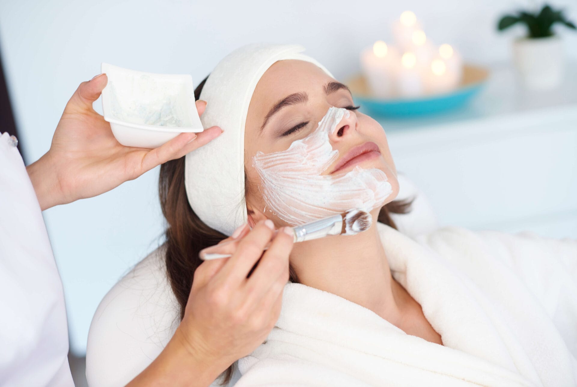 A woman in a robe, eyes closed, is in serene wedding prep mode, receiving a soothing facial treatment. As a gentle brush applies soft white cream to her skin, candles flicker gently in the blurred background.