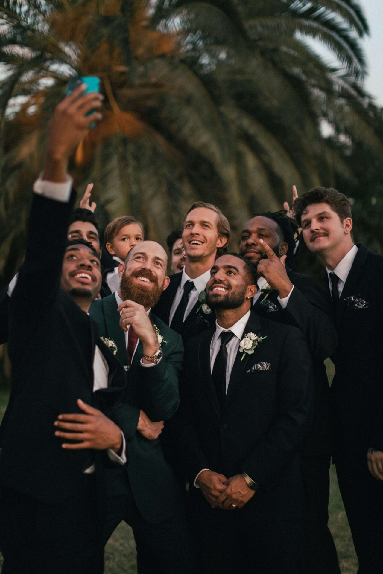 A group of people in formal attire pose for a selfie outside, capturing the vibrant scene with palm trees as their backdrop, while the excitement of wedding prep fills the air.