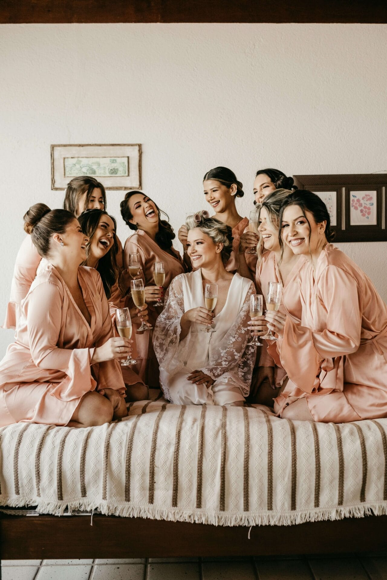 A bride in a white robe sits on a bed, surrounded by seven bridesmaids in pink robes, all smiles and clinking champagne glasses. The room buzzes with excitement as they toast to the perfect wedding prep.