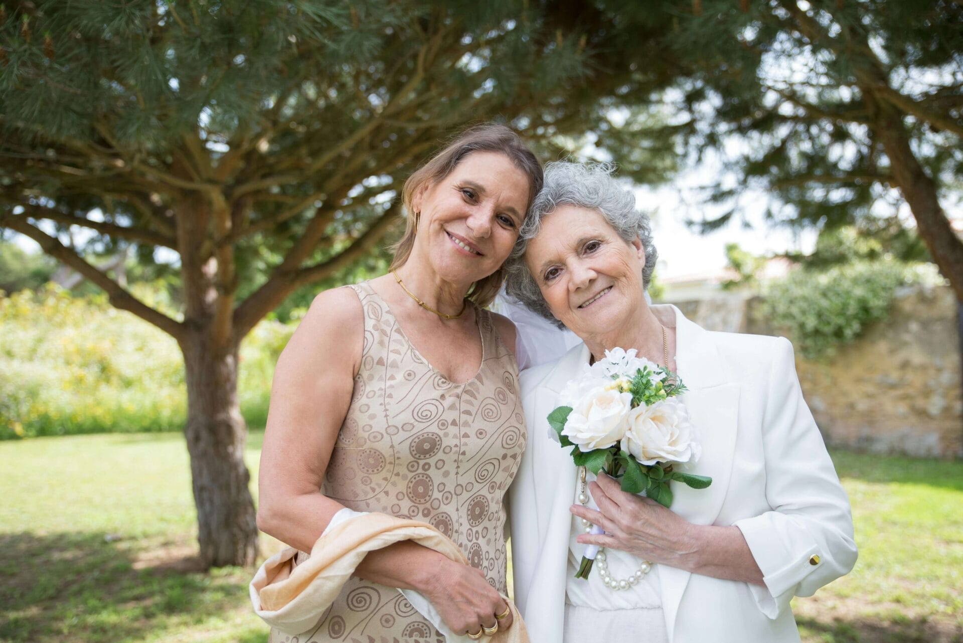 Two women standing together outdoors, one holding a bouquet of white flowers. They are smiling and standing under a large tree, perhaps savoring a serene moment amidst the joyous chaos of wedding prep.