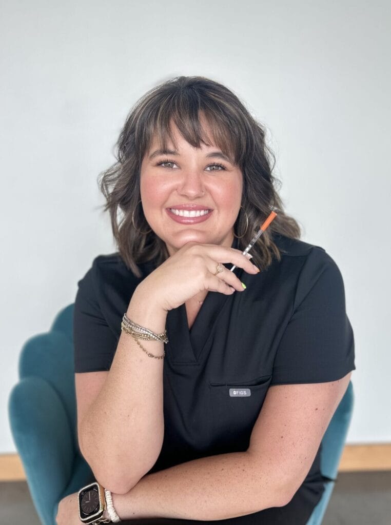 Woman with shoulder-length brown hair sits on a blue chair, smiling at the camera, wearing a black outfit and holding glasses in one hand.
